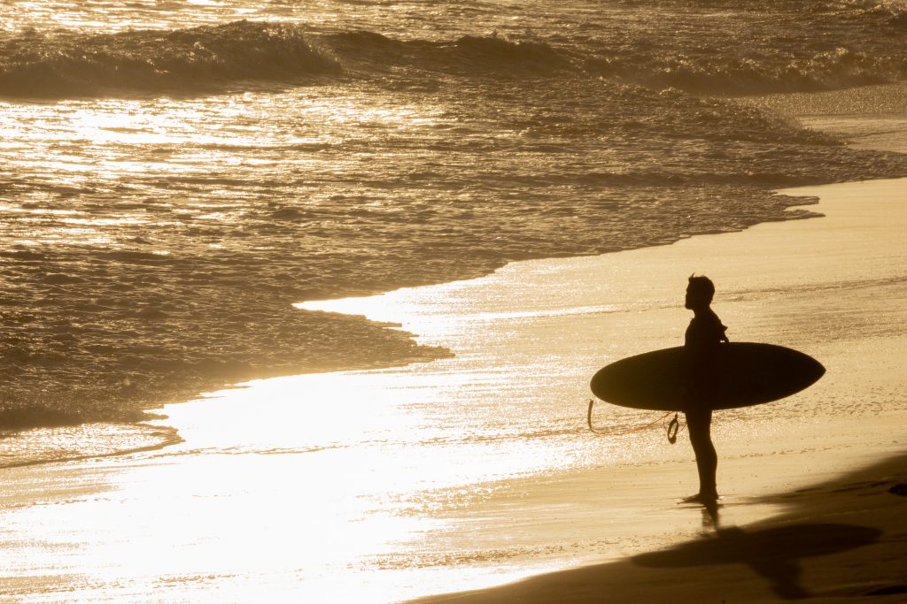 Silhouetted, isolated surfer standing on golden sand before sunw